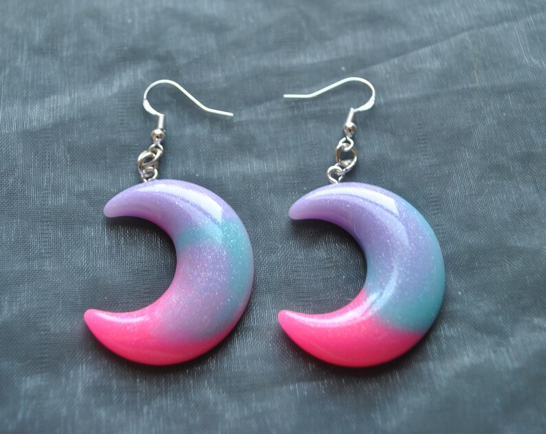 May include: A pair of crescent moon earrings with a pink and purple ombre effect and silver hooks.