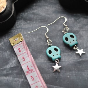 Puede incluir: Pendientes de calavera turquesa con ganchos plateados y colgantes de estrella. Los pendientes miden unos 4 cm de largo. Las calaveras tienen cuencas de ojos y nariz negras. Fondo gris.