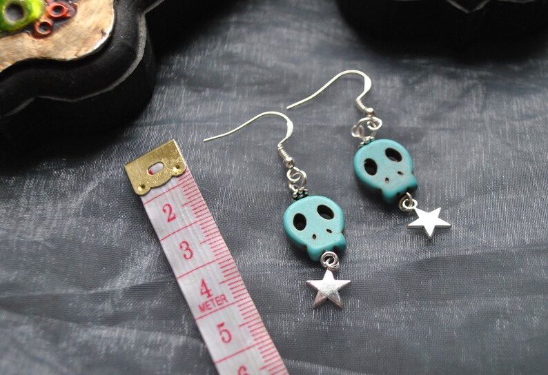 Puede incluir: Pendientes de calavera turquesa con ganchos plateados y colgantes de estrella. Los pendientes miden unos 4 cm de largo. Las calaveras tienen cuencas de ojos y nariz negras. Fondo gris.