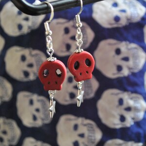 Puede incluir: Pendientes de calavera rojos con ganchos y detalles de cadena plateados. Cada pendiente presenta un peque&ntilde;o colgante de calavera roja y un dije de estrella plateada colgante. El fondo es una tela azul con patrones de calaveras blancas.