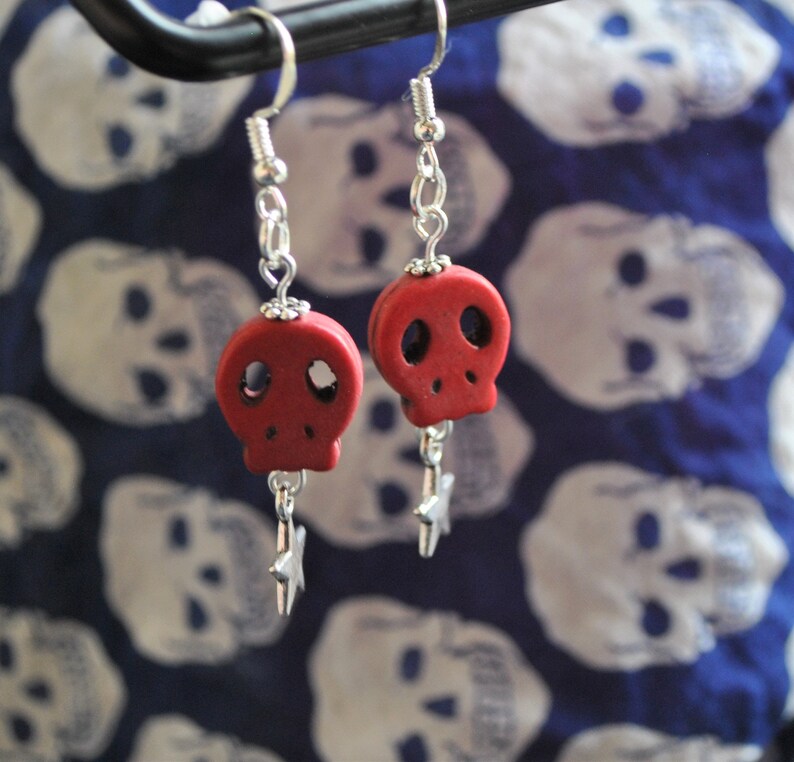 Puede incluir: Pendientes de calavera rojos con ganchos y detalles de cadena plateados. Cada pendiente presenta un peque&ntilde;o colgante de calavera roja y un dije de estrella plateada colgante. El fondo es una tela azul con patrones de calaveras blancas.