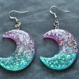May include: A pair of crescent moon shaped earrings with a pink and blue glitter gradient. The earrings are made of resin and have silver hooks.