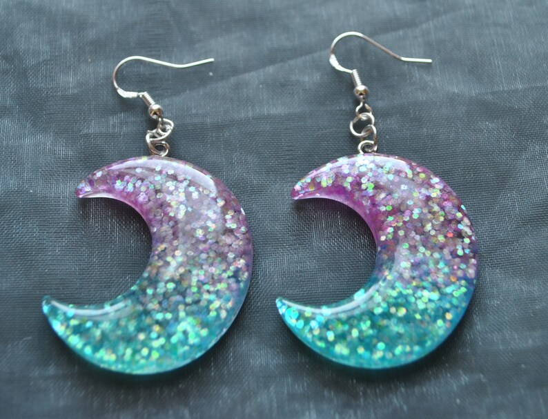 May include: A pair of crescent moon shaped earrings with a pink and blue glitter gradient. The earrings are made of resin and have silver hooks.