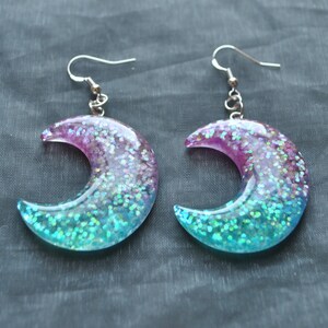 May include: A pair of crescent moon shaped earrings with iridescent glitter embedded in clear resin. The earrings are a gradient of pink, blue, and green.