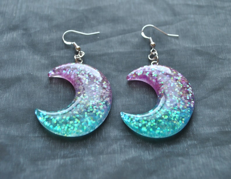 May include: A pair of crescent moon shaped earrings with iridescent glitter embedded in clear resin. The earrings are a gradient of pink, blue, and green.
