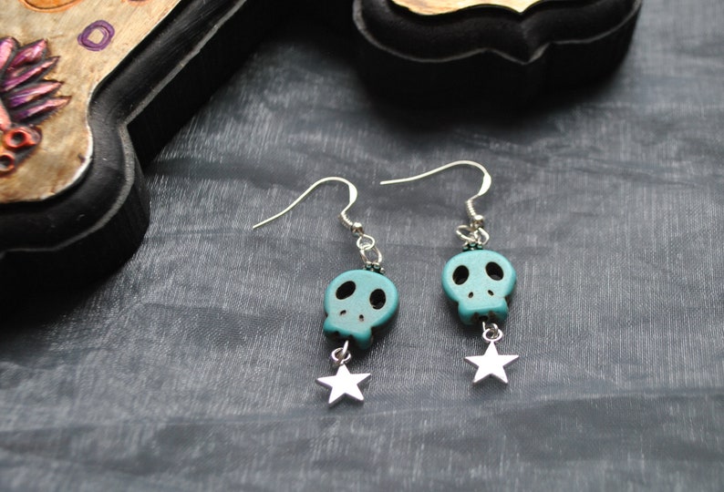 Puede incluir: Pendientes plateados con cuentas de calavera turquesa y colgantes de estrella. Los pendientes tienen cierre de gancho. La joya se exhibe sobre una superficie gris oscuro.
