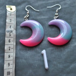 May include: A pair of crescent moon earrings with a pink, purple, and blue glitter finish. The earrings are made of resin and have silver hooks.