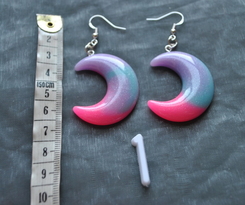 May include: A pair of crescent moon earrings with a pink, purple, and blue glitter finish. The earrings are made of resin and have silver hooks.