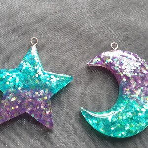 May include: Two resin charms, one shaped like a star and the other like a crescent moon. Both charms are filled with iridescent glitter and have a gradient of purple and teal colors.