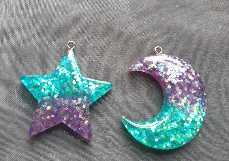 May include: Two resin charms, one shaped like a star and the other like a crescent moon. Both charms are filled with iridescent glitter and have a gradient of purple and teal colors.