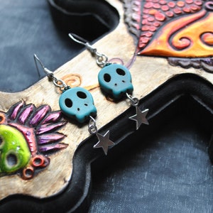 Puede incluir: Pendientes de calavera turquesa con colgantes de estrella plateada. Las cuentas de calavera tienen cuencas de ojos negras y est&aacute;n suspendidas de ganchos plateados. Los pendientes se exhiben sobre un fondo de madera tallada con dise&ntilde;os coloridos de calaveras y p&aacute;jaros.