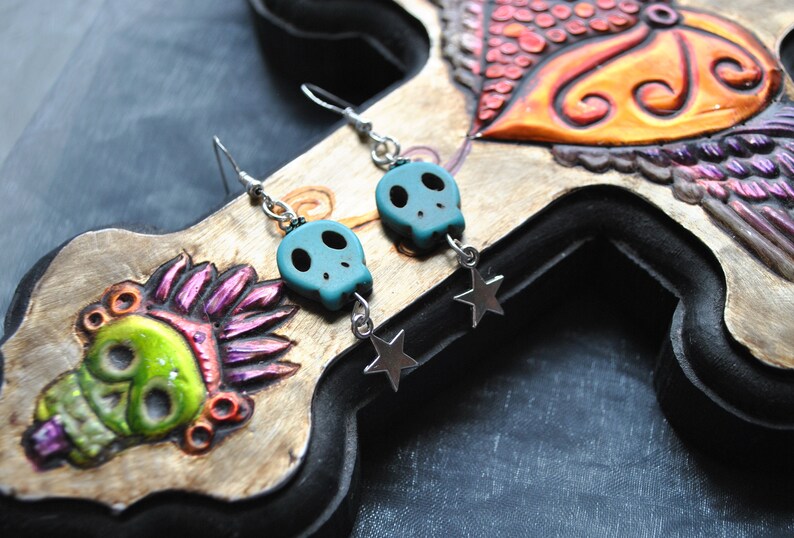 Puede incluir: Pendientes de calavera turquesa con colgantes de estrella plateada. Las cuentas de calavera tienen cuencas de ojos negras y est&aacute;n suspendidas de ganchos plateados. Los pendientes se exhiben sobre un fondo de madera tallada con dise&ntilde;os coloridos de calaveras y p&aacute;jaros.