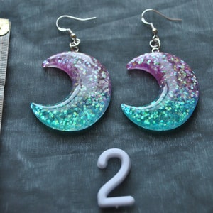 May include: A pair of crescent moon shaped earrings with a pink and blue glitter gradient. The earrings are made of resin and have silver hooks.