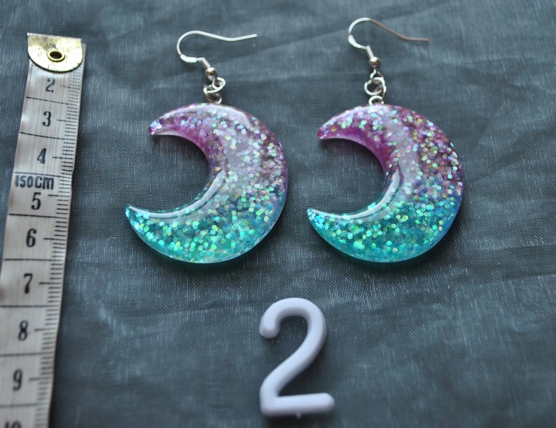 May include: A pair of crescent moon shaped earrings with a pink and blue glitter gradient. The earrings are made of resin and have silver hooks.