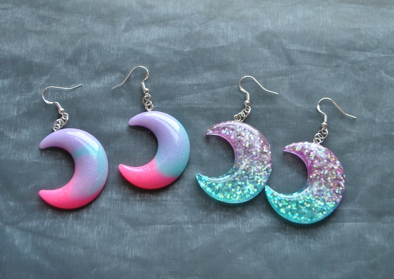 May include: A pair of crescent moon shaped earrings with glitter and ombre designs. One pair is pink and blue, the other is teal and purple.
