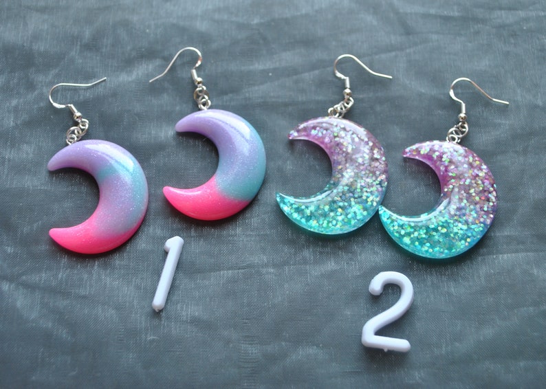 May include: A pair of crescent moon earrings with glitter embedded in the resin. The earrings are pink and blue with a gradient effect. The earrings are numbered 1 and 2.