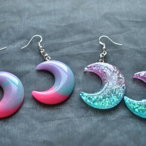 May include: A pair of crescent moon shaped earrings with iridescent glitter embedded in clear resin. The earrings are pink and blue with silver hooks.