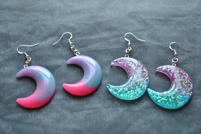 May include: A pair of crescent moon shaped earrings with iridescent glitter embedded in clear resin. The earrings are pink and blue with silver hooks.