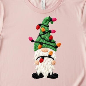 May include: A light pink t-shirt featuring a Christmas gnome design. The gnome has a green hat adorned with colorful Christmas lights, a white beard, and black shoes. The design is embroidered.