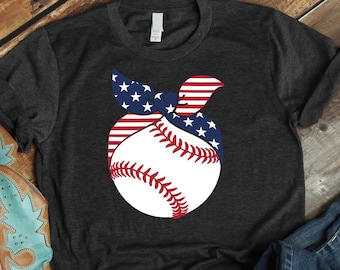 May include: A dark gray t-shirt with a white baseball graphic. The baseball is wrapped in a red, white, and blue bandana with stars. The shirt is laid out on a wooden surface with a pair of blue jeans and a brown cowboy boot.