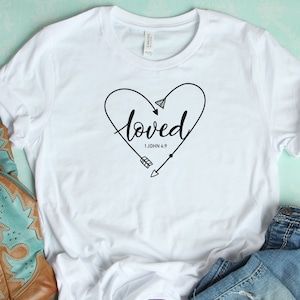 May include: White t-shirt with a black heart design and the word "loved" inside the heart. The heart has arrows on either side and the text "1 John 4:9" below the word "loved".