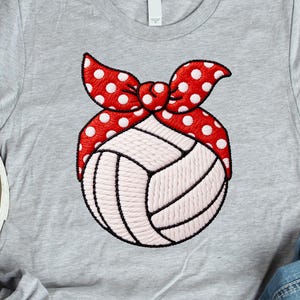 May include: A light gray t-shirt featuring a white volleyball with a red polka dot headband. The volleyball has black outlines and white stitching. The headband is red with white polka dots. The shirt is casual wear.