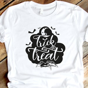 May include: White t-shirt with a black graphic design. The design features the words "Trick or Treat" in a stylized font, surrounded by bats, a spider web, a spider, and a crescent moon. The shirt is made of a soft material.