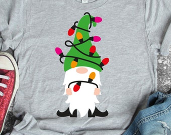 Könnte beinhalten: Ein graues T-Shirt mit einem grünen Gnom, der einen Hut mit rosa, orange und gelben Weihnachtslichtern trägt, die um ihn herumgewickelt sind.