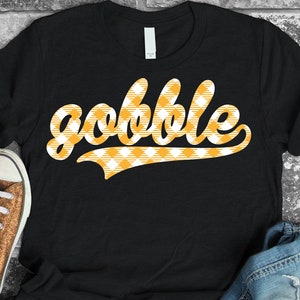 Puede incluir: Camiseta negra con un gráfico blanco "gobble" en un patrón de cuadros amarillo y blanco.
