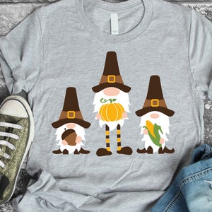 May include: A gray t-shirt with a design of three gnomes wearing pilgrim hats. The gnomes are holding a pumpkin, an acorn, and an ear of corn.