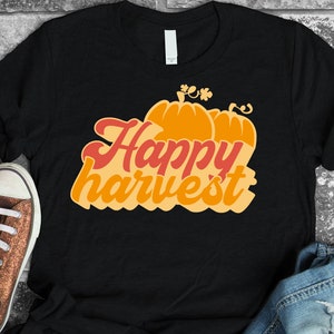 May include: Black t-shirt featuring a graphic of two orange pumpkins and the text "Happy Harvest" in red and orange.