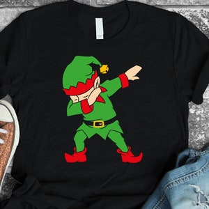 May include: Black t-shirt featuring a cartoon elf in a dabbing pose. The elf is dressed in green with red accents, a green hat with a gold bell, and red boots. The design is a fun, festive graphic.