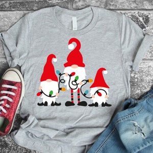 Puede incluir: Una camiseta gris con un diseño de tres gnomos sosteniendo luces de Navidad. Los gnomos llevan gorros rojos con pompones blancos y tienen barbas blancas. Las luces son rojas, verdes, azules y amarillas.
