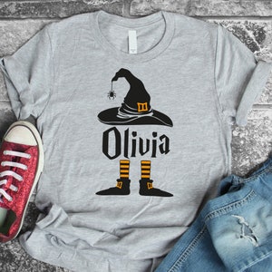 Puede incluir: Una camiseta gris con un sombrero de bruja negro, una araña y el nombre Olivia impreso en ella. El sombrero de bruja tiene rayas naranjas y una hebilla negra. Las piernas de la bruja son negras con rayas naranjas y zapatos negros.