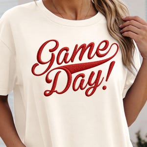 May include: Cream-colored t-shirt with the embroidered text "Game Day!" in a bold, red script. The design features a classic, sporty font, perfect for casual wear and game day events.