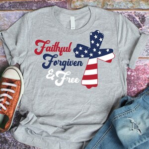 以下が含まれることがあります： グレーのTシャツに、赤、白、青のアメリカ国旗の十字架のデザインが施されています。Tシャツには「Faithful Forgiven & Free」という文字が白で印刷されています。