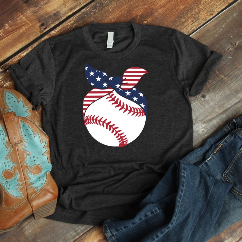 May include: A dark gray t-shirt with a white baseball graphic. The baseball is wrapped in a red, white, and blue bandana with stars. The shirt is laid out on a wooden surface with a pair of blue jeans and a brown cowboy boot.