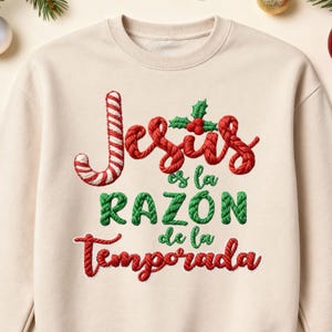May include: Cream-colored sweatshirt with the embroidered text "Jesus es la RAZÓN de la Temporada" in red, green, and white. The design includes a candy cane and holly leaves, perfect for the Christmas season.