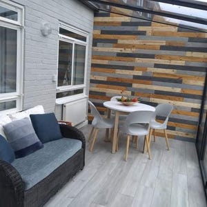 K&ouml;nnte beinhalten: Ein graues Korbsofa mit blauen Kissen steht in einem Wintergarten mit einem wei&szlig;en Holzboden. Ein wei&szlig;er Tisch mit vier wei&szlig;en St&uuml;hlen steht in der Mitte des Raumes. Die Wand hinter dem Tisch ist mit einem Holzpaneel aus recyceltem Holz verkleidet.