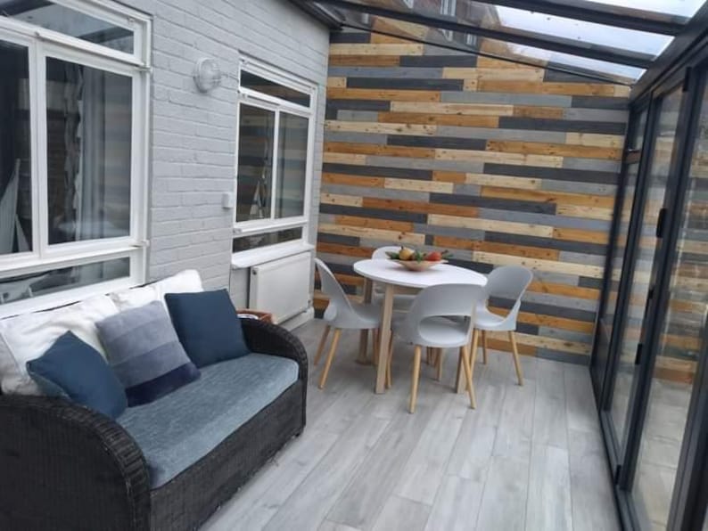 K&ouml;nnte beinhalten: Ein graues Korbsofa mit blauen Kissen steht in einem Wintergarten mit einem wei&szlig;en Holzboden. Ein wei&szlig;er Tisch mit vier wei&szlig;en St&uuml;hlen steht in der Mitte des Raumes. Die Wand hinter dem Tisch ist mit einem Holzpaneel aus recyceltem Holz verkleidet.