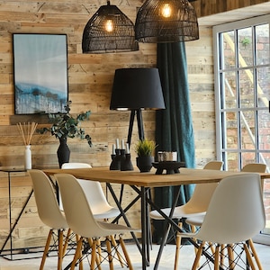 May include: A wooden dining table with black metal legs and six white chairs with wooden legs. The table is set with a vase of greenery, candles, and a decorative bowl. The room has a rustic wood wall with a large window and a black lamp with a black shade.