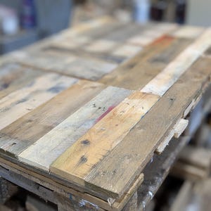 May include: A rustic wooden table top crafted from reclaimed pallet wood. The table top is constructed from various pieces of wood, each with a unique grain and colour. The wood is weathered and has a natural, rustic appearance.