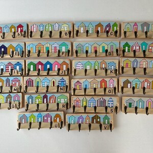 May include: A collection of twenty-two rectangular wooden wall hooks, each featuring a painted row of colourful beach huts. Every board includes four black metal hooks and a small wooden letter tile from A to V to identify each unique design.