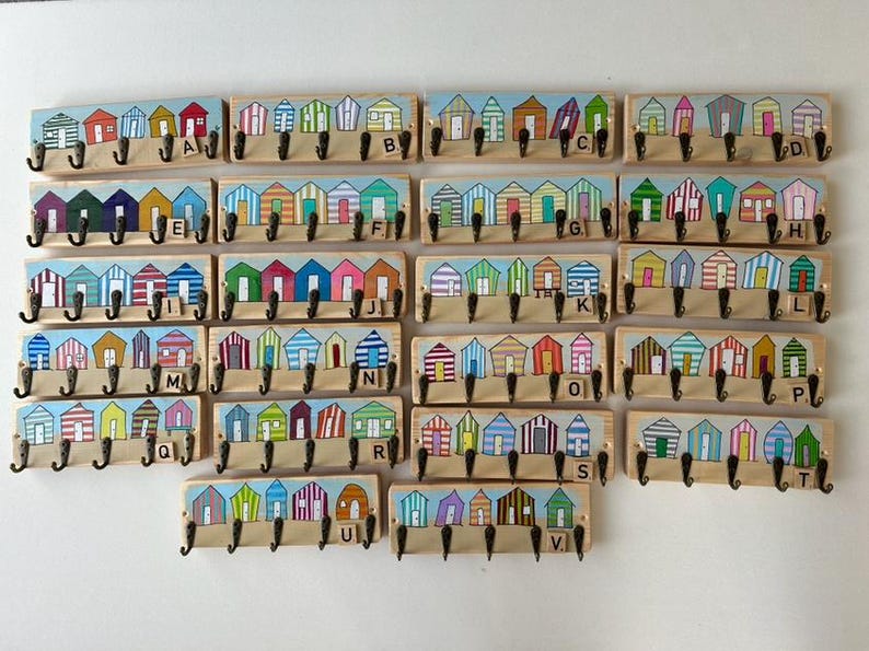 May include: A collection of twenty-two rectangular wooden wall hooks, each featuring a painted row of colourful beach huts. Every board includes four black metal hooks and a small wooden letter tile from A to V to identify each unique design.
