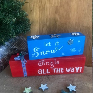 May include: Two wooden blocks stacked on top of each other. The top block is painted blue and says "let it snow" in white cursive writing. The bottom block is painted red and says "Jingle all the way!" in white cursive writing. There are several small wooden stars scattered around the blocks.