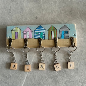May include: A rectangular wooden wall hook rack featuring a painted scene of five colourful beach huts. Five bronze-toned metal hooks are attached, each holding a silver keyring with a wooden tile displaying the letters B, E, A, C, and H.