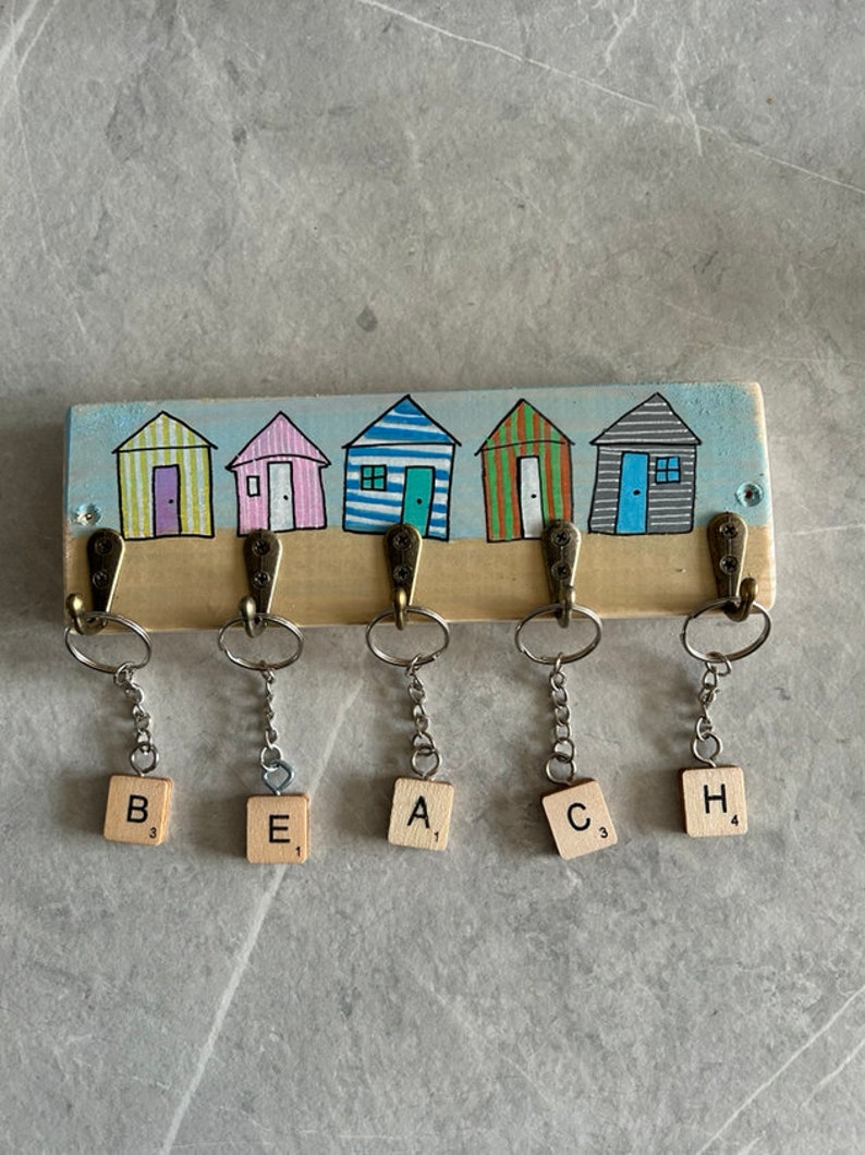 May include: A rectangular wooden wall hook rack featuring a painted scene of five colourful beach huts. Five bronze-toned metal hooks are attached, each holding a silver keyring with a wooden tile displaying the letters B, E, A, C, and H.