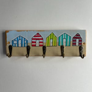 May include: A rectangular wooden wall hook rack featuring a hand-painted scene of five colourful beach huts in blue, red, lime green, teal, and grey stripes. The board includes five antique brass-toned metal hooks attached with visible screws.