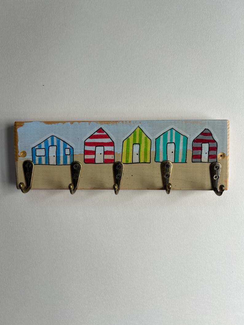 May include: A rectangular wooden wall hook rack featuring a hand-painted scene of five colourful beach huts in blue, red, lime green, teal, and grey stripes. The board includes five antique brass-toned metal hooks attached with visible screws.