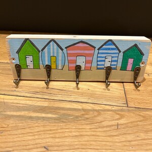 May include: A rectangular wooden wall hook rack featuring a hand-painted scene of five colourful beach huts in green, blue, and orange stripes. The piece includes five dark metal hooks attached with screws, set against a light blue sky and tan sand.